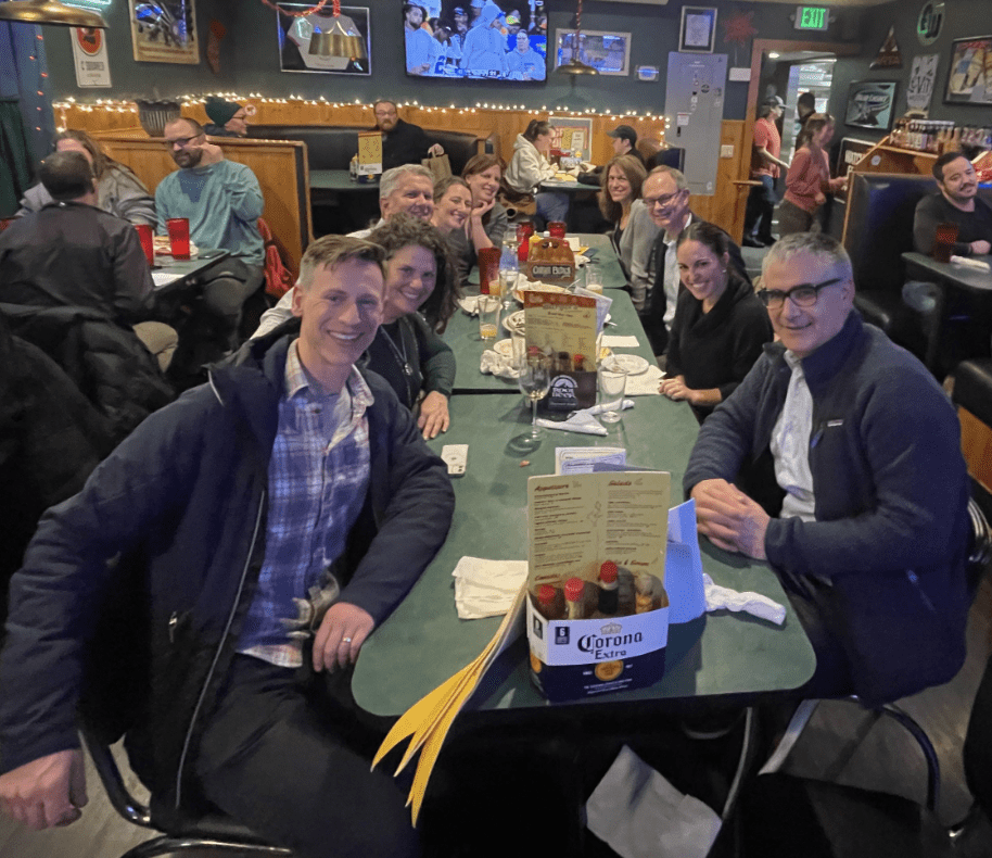 A group of people smiling and sitting around a table at a restaurant during a holiday happy hour event, with drinks and menus visible.
